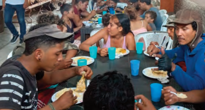 A group of people sit around a table, eating a meal together from plates and drinking from blue plastic cups.