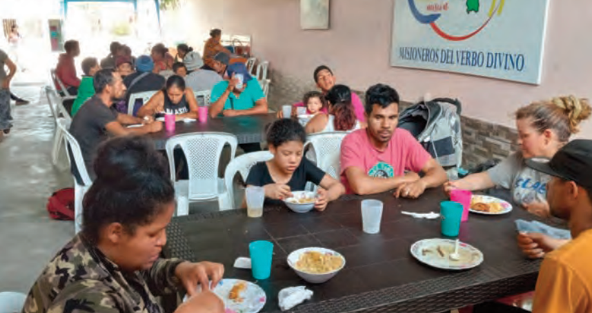 A group of people sit at tables eating food in a community dining area with white plastic chairs and cups on the tables.