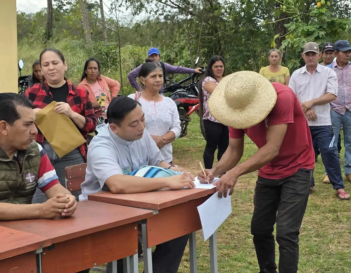 A man in a straw hat signs a paper at an outdoor table while others, seated and standing, watch in a rural setting with trees and grass.